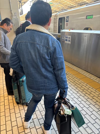 The image shows three men standing on a platform at a train station in Chiyoda City, Japan. The men are wearing casual clothes, including jeans and jackets. The man in the foreground is wearing a blue jean jacket and has a black bag slung over his shoulder. He is holding a teal rolling suitcase with one hand. The man in the middle is wearing a black jacket and has his back turned to the camera. The man in the background is wearing a gray jacket and has his back turned to the camera. The platform is made of brick and there is a train in the background. The train is white and has a number "14" on the side. The train is stopped at the platform and the doors are open. The image is taken at the station located at 35.682 latitude and 139.768 longitude.