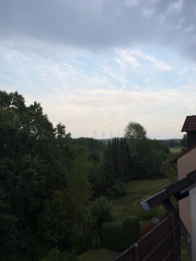 A wide-angle, high-angle shot depicts a scenic view from a balcony in Creußen, Germany. The foreground is dominated by a dark brown wooden railing and a portion of a light-colored building with a terracotta-tiled roof. Beyond the railing, a lush green lawn stretches out, bordered by a wooden fence and dense trees. In the middle ground, a line of various trees, including coniferous and deciduous varieties, creates a natural barrier. In the background, a field with some scattered trees is visible, and on the horizon, several wind turbines stand tall against a cloudy, pale blue sky. A faint contrail is visible in the sky. The overall scene suggests a peaceful, rural or semi-rural setting with a focus on natural elements and renewable energy sources.