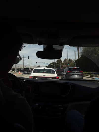 The image depicts a view from inside a car in La Quinta, United States. The driver, wearing sunglasses, is behind the wheel of a Chrysler Pacifica with a black dashboard. Outside the windshield, there's a white Kia and a gray Hyundai Tucson, among other cars stopped at a traffic light on a street with telephone poles and palm trees. The sky is clear and lightly clouded.
