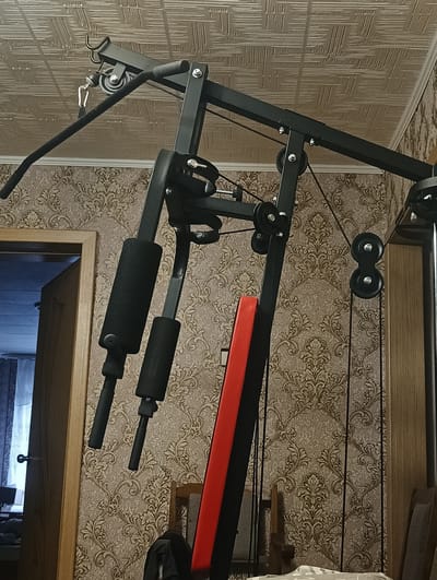 This is a medium shot, eye-level, indoor, full-color photograph of a home gym setup in what appears to be a residential room. The room has patterned wallpaper and a textured ceiling. The main subject is a black metal multi-gym apparatus, partially visible. The gym equipment features various pulleys, cables, and padded attachments. A red and black padded bench is also visible, leaning against the gym structure. To the left, a partially open wooden door reveals a glimpse of another room with a window and curtains. No people are present in the image. The location is identified as Тайжина, Russia.