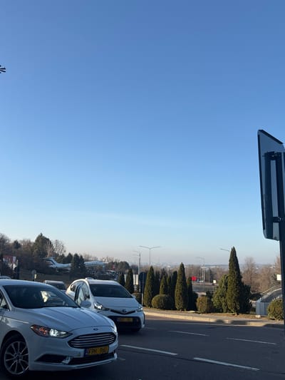 The image presents a daylight outdoor scene on a clear, sunny day in the vicinity of Chișinău International Airport, which borders the commune of Bubuieci, Moldova.

In the foreground, an asphalt roadway is visible with a partial white dividing line. Two white vehicles are present: a Ford Fusion sedan occupies the lower-left, displaying a Moldovan license plate "FPH 960" with yellow characters. Immediately behind it and slightly to the right, a white Toyota Prius or similar hybrid vehicle is partially visible, bearing the Moldovan license plate "AOT 01". Both vehicles face slightly right. On the far right, a dark, reflective, rectangular object, possibly part of a sign structure, is partially cropped.

The midground features a landscaped area with a mix of evergreen trees (conical and rounded shapes) and bare deciduous trees, suggesting a non-summer season. Prominently positioned on an elevated, grassy, and tree-covered mound is a white Tupolev Tu-134 aircraft, serving as a static monument. Streetlights are visible further along the road.

The background shows a distant, hazy urban or industrial landscape with various buildings and structures, all under a vast, cloudless blue sky that dominates the upper portion of the frame. No individuals are visible, and no active interactions or specific actions are discernible beyond the presence of parked or slowly moving vehicles.