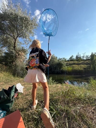 A young girl with blonde hair stands in a grassy field, holding a blue fishing net above her head. She is wearing a black t-shirt with a colorful design on the back and a white skirt. In the background, there is a body of water with trees and a clear blue sky with some clouds. There are two fishing rods in the water, and a person in the water wearing a red shirt. A green backpack and an orange mat are on the ground near the girl. The scene appears to be a rural area, possibly on the outskirts of Пушкинское сельское поселение, Ukraine.