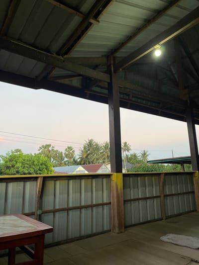 The image is taken from inside a covered structure with a corrugated metal roof supported by wooden beams and posts. A single light bulb hangs from the ceiling. In the foreground, a wooden table with a dark red frame and a lighter surface is visible. The structure opens to an outdoor scene. Beyond a corrugated metal wall, a line of lush green trees and palm trees is visible. Several houses with colorful roofs are nestled among the foliage. The sky above is a pale gradient of pink and blue, suggesting either dawn or dusk. The environment appears to be a residential area, possibly in a tropical region given the vegetation. The specific location is the city of Turumame, Indonesia.
