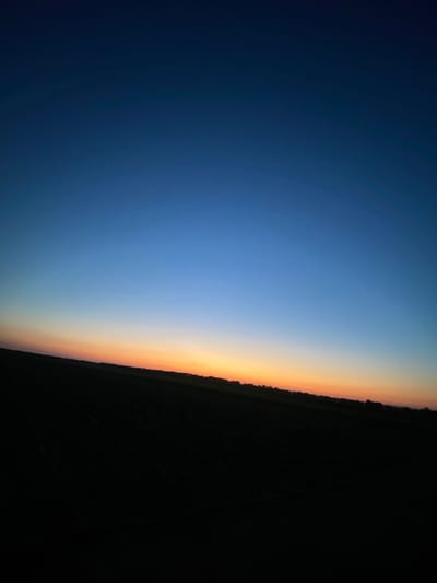 The image captures a twilight scene over a field in Luliakovo, Bulgaria. The sky transitions from a deep navy blue at the top to a vibrant orange and yellow gradient near the horizon. The lower portion of the image is dominated by a dark, silhouetted landscape, likely a field, with a faint line of trees or vegetation along the horizon. The overall impression is one of peaceful solitude as day turns into night.
