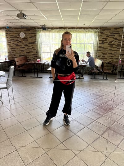 A woman stands in what appears to be a dining area or cafe, taking a mirror selfie. She is wearing a black sweatshirt with white lettering, black pants, and a red fanny pack. Her hair is blonde and pulled back. She is holding a pink smartphone in her right hand.

The environment has a tiled floor and walls covered with a faux stone pattern. There are tables and benches set up in the background, some of which are partially obscured by sheer curtains. A clock is visible on the wall. In the background, a man is seated at a table, looking down and appearing to be eating or working on something. The scene is indoors with natural light coming from the windows. The location is identified as Глебычево, Russia.