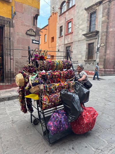 A vendor sits behind a flower crown display on a wheeled cart on a San Miguel de Allende street. The vendor, an older woman with gray hair, is wearing a black and white floral shirt. She is leaning over the cart, tending to the wares. The cart is loaded with flower crowns of various colors and styles hanging on metal rods. Above the crowns are bags of items, including a red floral bag, black plastic bags, and a purple patterned bag. The street is made of gray stone. A person in a gray shirt and jeans is walking in the background. Buildings line the street, with one having a sign labeled "Canal Manzana No. 14" with a downward-pointing arrow. The buildings are constructed of stone and are painted in shades of gray, orange, and yellow.
