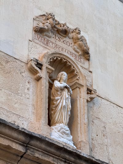 The image captures a stone sculpture of a reclining figure, possibly depicting the Virgin Mary, located on a wall in Vinaròs, Spain. The sculpture is set within a decorative stone frame. To the left of the figure are relief carvings, including angels and inscriptions reading "VENICORONABERIS I.S.60.". The surrounding wall is of a light-colored stone material. The sculpture is likely part of an architectural element of a church or historical building.
