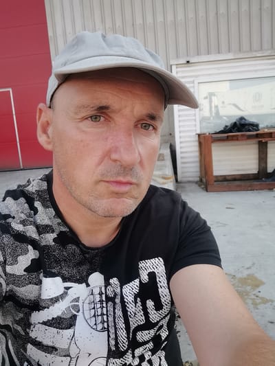 The image shows a man wearing a light gray baseball cap and a black t-shirt with a white graphic print. He is looking directly at the camera with a neutral expression. The background consists of a building with a red panel on the left and a white corrugated metal wall on the right, with a window that shows an interior scene. There is also a wooden structure with some black plastic bags on it. The ground appears to be concrete, with some patches of wetness and debris.
The man's features include short, dark hair, light-colored eyes, and some stubble on his face. His t-shirt has a complex design that includes what looks like a skeleton or a large animal. The setting appears to be outdoors, possibly near a warehouse or industrial building, during daylight. The lighting suggests it might be late afternoon or early morning due to the soft shadows. The overall mood is casual and candid. No text is clearly legible in the image, though the graphic on the shirt may contain text that is not discernible. The location context provided is Slanchevo, Bulgaria.