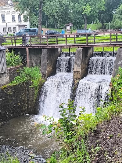This image captures a view of the Venta Rapid in Kuldīga, Latvia, also known as the Venta waterfall. The waterfall is composed of several channels of water cascading down a concrete structure. A wooden pedestrian bridge spans across the top of the waterfall structure, with two individuals standing on it, looking towards the water. Beyond the bridge, a dark-colored van is parked, and in the distance, several other vehicles and a white building with a distinctive architectural style are visible. The scene is framed by lush green trees and vegetation, with grassy slopes on the right side of the image. The water in the foreground pool below the falls appears murky.