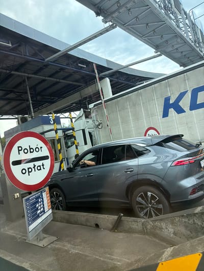 The image shows a gray SUV at a toll booth on a cloudy day. The toll booth is lane number 3, indicated by a blue sign. A red and white circular sign with Polish text "Pobór opłat" which translates to "Payment collection" is visible in the foreground. A price list for different vehicle categories is also displayed. A truck is visible behind the SUV. The background shows a large building with "KC" logo on it. The overall scene is a toll collection point in Mysłowice, Poland.
