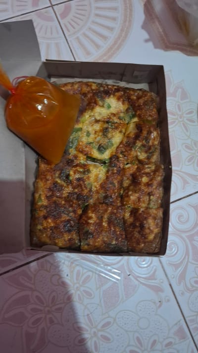 This media file displays a close-up, overhead shot of an Indonesian savory omelet, possibly martabak telur, cut into rectangular pieces and presented in a brown cardboard box. The omelet has a rich, golden-brown, slightly crispy exterior with visible green scallions or chives mixed throughout its eggy filling. Adjacent to the omelet, a clear plastic pouch filled with a vibrant orange sauce, likely a sweet and spicy dipping sauce, is positioned on the left side of the box.
The food is set against a backdrop of a patterned tiled surface, featuring a repeating floral design in shades of pink and white. The tiles suggest an indoor setting, likely a home or a food stall. The lighting appears to be natural or soft indoor lighting, casting subtle shadows. There are no people or specific weather cues visible. The overall impression is one of a prepared meal, ready to be served or consumed. The location context of Batam, Indonesia, further grounds the dish within its cultural culinary tradition. No visible text is present in the image.