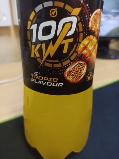 A bottle of "100 KWT Tropic Flavour" energy drink is positioned on a desk in Shakhunya, Russia. The bottle, predominantly black and yellow, features a graphic of a stylized "100 KWT" logo with lightning bolt elements, and images of sliced citrus fruits. The drink inside is a bright yellow. The desk surface appears to be a dark mat. In the background, a white power outlet with a green indicator light is visible on a wall, and a blurred electronic screen or monitor is also present. The lighting suggests an indoor environment. There are no people visible in the image.