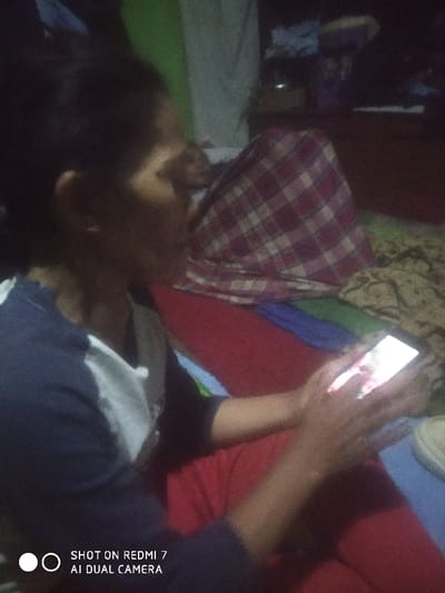 The image depicts an indoor scene in Lamajang, Indonesia, under dim lighting. In the foreground, a person is seated or reclining, visible from the chest upwards in a right-profile view. This individual is wearing a dark blue and white long-sleeved shirt and is actively holding a white-backed smartphone with both hands. The smartphone screen is illuminated, displaying an image that appears to be a person or a face. The person's lower body is covered by a red fabric, possibly pants or a blanket.

In the mid-ground, behind the foreground individual, another person is partially visible, largely covered by a red and white plaid blanket. This individual appears to be reclining or lying down. Various textile layers are present in the immediate environment, including a red fabric beneath the foreground person, a blue fabric layer, and a beige-patterned fabric on the right side of the frame. The background is dark and indistinct, showing vague outlines of furniture or shelves containing various dark objects. A vertical green surface is partially visible on the far left. A watermark stating "SHOT ON REDMI 7 AI DUAL CAMERA" is present in the lower-left corner of the image.