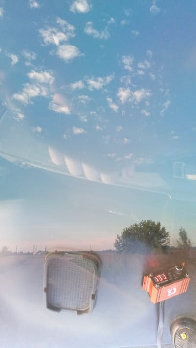 The image is a reflection on a shiny, light blue surface, likely a car's dashboard or hood. The reflection shows a bright blue sky with scattered white clouds. In the reflection of the sky, there are curved, wavy white shapes that appear to be reflections of some external objects.
In the lower portion of the reflection, a landscape is visible. This landscape appears to be an outdoor scene with a flat, light-colored ground, possibly a road or a field. In the distance, there are power lines or industrial structures. A few trees are also visible on the right side of the reflected landscape.
On the actual surface of the blue object, in the foreground, there is a black, rectangular object with a grid pattern, possibly a speaker grill or a vent. To the right of this, there is a small, orange electronic device with a digital display showing "13.3" in red LED digits. This device has a dial and some buttons on its top surface, and it has ridged sides. A dark-colored shoe with a yellow logo is visible in the bottom right corner of the image, partially on the blue surface.
The image is taken with a low angle, suggesting the camera was positioned close to the blue surface. The lighting is bright, indicating daylight. The location of Zuy, Russia is not explicitly discernible from the visual elements of the image itself.