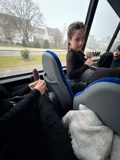 Inside a vehicle, a young person in a dark top sits near the window, looking at the camera. Outside, houses and bare trees are visible on a gray day. A light gray seat is in front, with a beige sweater draped over it. In the foreground, someone is holding a finger puppet near the window. Another person is visible in the upper part of the frame.