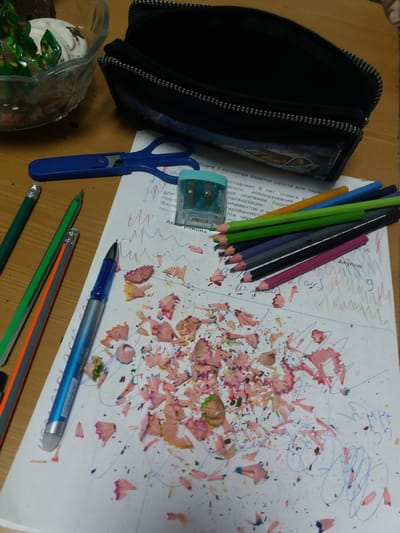 A desk in what appears to be a home is covered with drawing and writing implements, along with a variety of other objects. At the left, a black pencil case lies open. A clear plastic container with green objects is located partially in the lower-left corner. To the right of the pencil case is a blue pencil sharpener, and next to that, a blue folding pair of scissors. A number of colored pencils are laid out; they include colors such as blue, green, yellow, black, pink, and grey. A few regular, wooden pencils are also there. One blue and silver pen is near the center of the composition. The scene also features a sheet of paper which has notes written in Russian, scribbles, and pink pencil shavings. This image was taken in Бородулиха ауылдық округі, Kazakhstan, as indicated by the Russian language text on the paper.