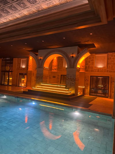 The image depicts a courtyard in Ürgüp, Turkey, featuring a pool and a building with arched doorways and illuminated staircases. The building's architecture includes brickwork and decorative patterns. The pool's surface reflects the building's lights and architectural details. No people are visible in the scene.