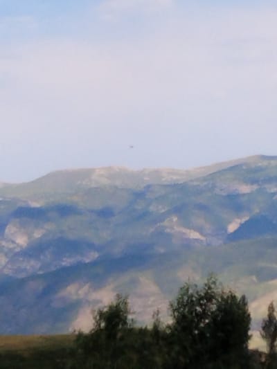 The image depicts a distant view of rolling hills and mountains under a hazy, light blue sky. The landscape appears to be sparsely vegetated, with shades of green, brown, and blue dominating the scene. In the foreground, the tops of dark green trees are visible, suggesting a natural, possibly rural or mountainous, setting. There is a small, dark, indistinct object visible in the sky above the distant mountains, too small and blurry to identify. The overall impression is one of a vast, open natural landscape with a clear sky. The location is identified as Ак-Дөбө, Kyrgyzstan.