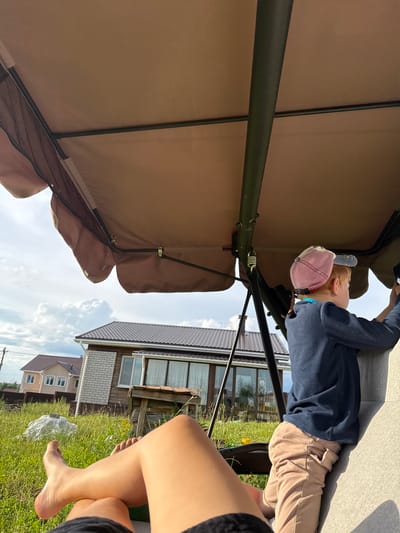 The image shows two people relaxing outdoors. One person, a child with red hair wearing a pink cap and a navy blue sweater, is lying down under a tan canopy, possibly a patio umbrella or swing. The child's head is resting on a pillow. The other person's legs are visible in the foreground, suggesting they are sitting or lying beside the child. The setting appears to be a backyard with a grassy area, some vegetation, and houses in the background. The sky is partly cloudy, indicating fair weather. The overall mood seems relaxed and peaceful, suggesting a casual outdoor leisure activity on a sunny day. The scene is likely in Пычанки, Russia, as indicated by the location context. There is no visible text in the image.