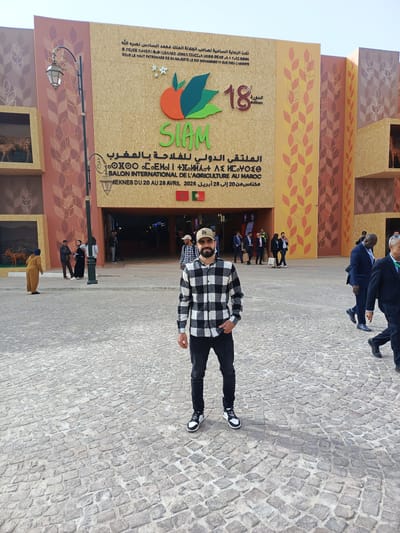 The image shows an outdoor scene at the entrance of a large exhibition building, located at Al Machouar – Stinia, Morocco. In the foreground, a man with a beard, wearing a black and white plaid shirt, black jeans, and a beige baseball cap with an "LA" logo, stands facing the camera. He has one hand in his pocket.
The background is dominated by the facade of a building, featuring decorative panels in shades of yellow, orange, and brown with stylized wheat stalk patterns. Display boxes on the sides of the entrance showcase animal figurines, such as a cheetah or deer. A traditional street lamp is visible on the left.
The building's signage identifies the event as the "SALON INTERNATIONAL DE L'AGRICULTURE AU MAROC" (International Agriculture Show in Morocco), also known as "SIAM." The SIAMS logo, depicting fruits and leaves, is prominent. The text further states it is the "18ème édition" (18th edition), held "SOUS LE HAUT PATRONAGE DE SA MAJESTÉ LE ROI MOHAMMED VI QUE DIEU L'ASSISTE" (Under the High Patronage of His Majesty King Mohammed VI, may God assist him). The event dates are "MEKNES DU 20 AU 28 AVRIL 2026." Moroccan and Portuguese flags are displayed above the entrance.
Various people are seen in the mid-ground and background, walking in and out of the building, suggesting an active event. Some are dressed casually, while others are in business attire. The ground is paved with cobblestones. The lighting is bright and clear, indicating a sunny daytime.