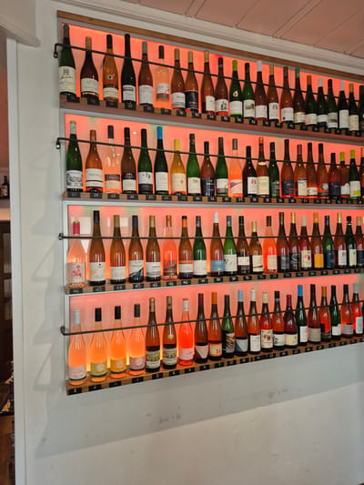 This image captures a wall-mounted wine display within a commercial establishment in Bernkastel-Kues, Germany. The display consists of six horizontal shelves, each supported by two parallel metal rods, against a plain white wall. Each shelf is backlit with a warm, reddish-orange light that illuminates the wine bottles and enhances the visibility of their contents.
A total of 92 wine bottles are displayed, sequentially numbered on small, light-colored wooden blocks positioned directly beneath each bottle. The numbering begins with '1' on the bottom-right of the lowest shelf and ascends to '92' on the top-left of the highest shelf.
The bottle count per shelf, from top to bottom, is as follows:
* Top shelf: 9 bottles (numbered 92 to 84)
* Second shelf: 18 bottles (numbered 83 to 66)
* Third shelf: 18 bottles (numbered 65 to 48)
* Fourth shelf: 18 bottles (numbered 47 to 30)
* Fifth shelf: 18 bottles (numbered 29 to 12)
* Bottom shelf: 11 bottles (numbered 11 to 1)
The bottles vary in glass color, including dark green, dark brown, and clear, and contain wines exhibiting a spectrum of colors from deep golden amber to various shades of pink (rosé) and light yellow. Many labels are visible, displaying German text and identifiable vintner names such as 'Müller Thurgau,' 'Weber,' 'Markus Molitor,' 'PaZen,' 'Sebastian Erbeldinger,' 'Leitsch,' 'Bollig,' 'Kitzmuhle,' and 'Pichter.' These labels indicate the German origin of the wines, typical of the Bernkastel-Kues region.
The surrounding environment includes a clean, white wall surface. Above the display, a white ceiling with visible paneling is present. To the far left, a vertical white architectural element, likely a door frame or wall corner, is visible. A dark, out-of-focus wooden object, possibly a shelf or cabinet, is partially visible in the lower left background. No individuals are present in the image.