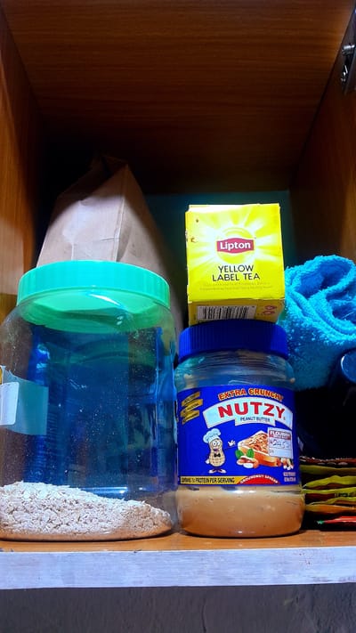 This media file captures a close-up, rotated view inside a wooden cupboard or shelf, likely in a kitchen or pantry in Umuigbekwo, Nigeria. The image is oriented sideways, with objects appearing to lie on their sides.
The main subjects are various food items and a cleaning cloth. Prominently featured is a bright yellow box of Lipton Yellow Label Tea, which shows some wear. Text on the box includes "Lipton", "YELLOW LABEL TEA", and a health claim: "Tea is a rich source of flavonoids. Dietary flavonoids may help maintain a healthy heart."
Next to the tea box is a blue-lidded jar of "NUTZY EXTRA CRUNCHY PEANUT BUTTER." The label specifies "PEANUT BUTTER," "GROUNDNUT SPREAD," and "CONTAINS 7g PROTEIN PER SERVING." Other notable text on the label includes "FLOURISH MEGA MAN," "N2450," "S/N," "HEALTHY," "NO CHOLESTEROL," and "NO PRESERVATIVES." The "N2450" likely indicates a price in Nigerian Naira, reinforcing the specified location context.
Below the peanut butter jar is a large clear plastic jar with a bright green lid, filled with what appears to be oatmeal or rolled oats. Behind the tea and peanut butter, a crumpled blue microfiber towel or cloth is visible. In the lower left, part of a brown paper bag is tucked away, and behind the items on the right, several small, colorful packets or sachets can be seen.
The setting is dimly lit, suggesting an interior space away from direct light, with the wooden surfaces of the cupboard creating a warm, enclosed environment. No specific activity or event is taking place; the image is a still life of stored provisions.