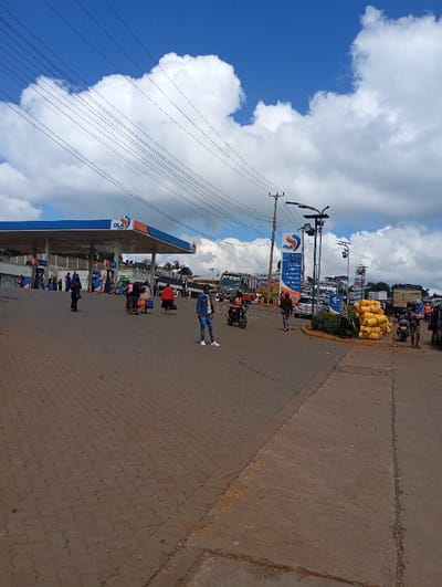 An Ola gas station is visible under a cloudy blue sky. Power lines stretch across the upper portion of the image. The foreground features a brick-paved area with people and motorcycles. Further back, there's an active area with vehicles, buses, and individuals around the gas station. Yellow containers are stacked near a circular barrier.