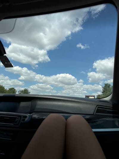 The image shows a view from inside a car looking out through the windshield. The sky is a bright blue with scattered white cumulus clouds. In the distance, there are trees and a small structure that appears to be a building or a truck. The dashboard of the car is visible in the foreground, and a person's legs are also visible, suggesting the photo was taken from the passenger seat. The scene appears to be set during the daytime, with clear weather. There is no visible indication of the specific location in Poysdorf, Austria.