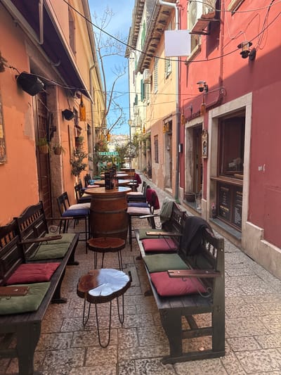The image depicts a narrow street in Grad Rovinj, Croatia, lined with buildings of varying heights and colors, predominantly reddish-orange hues. Tables and chairs occupy the street, suggesting an outdoor dining area, potentially for a restaurant or cafe. Some tables are constructed from wooden barrels and others are made of tree trunks. Seating consists of wooden benches with cushions. The buildings display architectural details such as windows, doors, and decorative elements. Wires and lights are strung across the street. The sky is visible in the upper portion of the frame.