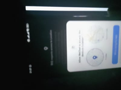 The image shows a smartphone screen displaying a WeChat location permission request. The request asks to "Allow WeChat to access this device's location." There are two circular icons, one with a map-like graphic and another with a blue map marker. A blue button with the text "Allow" is visible on the right side of the screen. The phone is held at an angle, and the background is dark. The overall scene is consistent with a user interacting with a mobile application. There is no identifiable information or context related to the city of Azume, Nigeria.