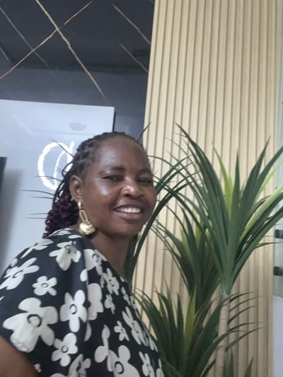 A woman is smiling at the camera. She is wearing a black and white floral dress and gold earrings. Behind her is a plant and a wall with horizontal stripes. A dark grey wall with gold lines is visible in the background. This image cannot be geographically located in Mopo-Akinlade, Nigeria.