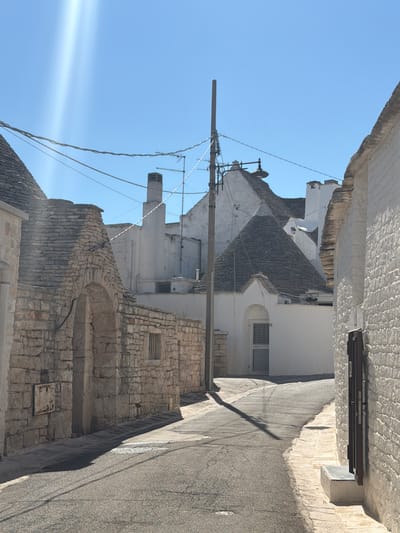 This image depicts a narrow, paved street in Alberobello, Italy, lined with traditional whitewashed buildings known as "trulli." The architecture features distinctive conical stone roofs. On the left side of the street, a section of a trullo with a stone archway and rough stone walls is visible. Further down the street, a white building with a large, conical grey stone roof is prominent. The right side of the street is dominated by a whitewashed building with a textured stone facade and a dark wooden door. Overhead, power lines and a utility pole are strung across the clear blue sky. The sun is shining brightly, casting sharp shadows across the street and the buildings. The scene is devoid of people, creating a tranquil and historic ambiance.