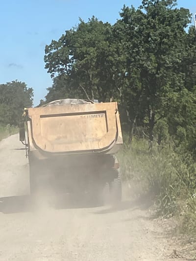 A yellow dump truck is driving down a dirt road, kicking up a cloud of dust. The truck is loaded with sand. The road is lined with trees and vegetation. The sky is blue and there are a few clouds. The scene appears to be in a rural area, possibly outside of Medovo, Bulgaria.