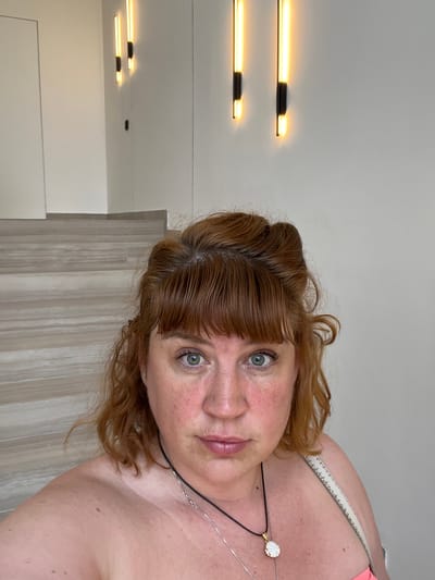 A woman with reddish-blonde hair and green eyes is taking a selfie in an indoor environment that appears to be a staircase in a modern building. The background features a neutral-colored wall with two vertical strip lights illuminating the area. To the left, there are steps of a staircase with a wood-like texture. The woman is wearing necklaces, and her shoulders and upper chest are visible, suggesting she is wearing an off-the-shoulder top. The lighting in the image is bright, and there are no other people visible. The location is described as Pitsunda, Abkhazia.