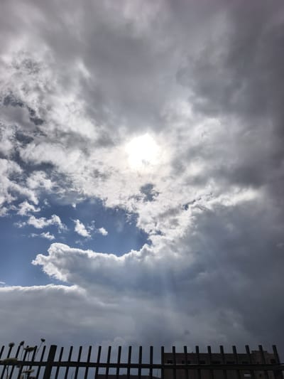 The image captures a cloudy sky over the Jezreel Valley Regional Council, Israel. The sun is visible through a break in the clouds, with light rays streaming down. The sky transitions from light blue with white clouds to dark gray, suggesting an approaching storm. In the foreground, there is a wooden fence with plants and a building behind it.