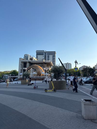 The image depicts a public square with a modern architectural building in the background. The building has a unique rounded wooden structure integrated into its facade, featuring large windows and balconies. Several people are walking and riding electric scooters in the square. Some individuals are dressed casually, and the overall atmosphere suggests a warm day. In the background, there are other buildings, trees, and parked cars, indicating an urban setting. A pedestrian crossing sign is visible, suggesting a street adjacent to the square. The sky is clear blue, indicating good weather. The foreground features a patterned paved surface with a concrete planter and a trash receptacle. The image appears to be taken during daylight hours, likely late afternoon or early evening, given the lighting. The presence of multiple electric scooters suggests a contemporary urban environment where such modes of transport are popular.