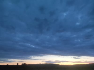 The image depicts a wide shot of a landscape under a dark, cloudy sky. The horizon is marked by a low, silhouetted line of trees and hills. The sky is dominated by heavy, dark blue clouds, with hints of lighter, warmer colors, such as yellow and orange, visible near the horizon, suggesting either dawn or dusk. The foreground consists of a dark, indistinct grassy area. There are no people or animals visible in the image. The location is Cherevky, Ukraine.