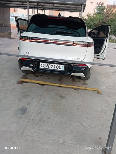 The image shows the rear view of a white SUV parked outdoors under a covered structure. The car's doors are open. The license plate is visible and reads "10W0210W". In the background, a building with the sign "LOR" is visible, along with trees and another building with signs that read "TERAPEVT" and "GINEX". The ground is concrete and appears wet in places. There is a yellow pipe lying on the ground in front of the car. The overall scene suggests the car is at a car wash or service area in an urban setting, likely in Bekabad, Uzbekistan, given the context provided. No people are visible in the image.