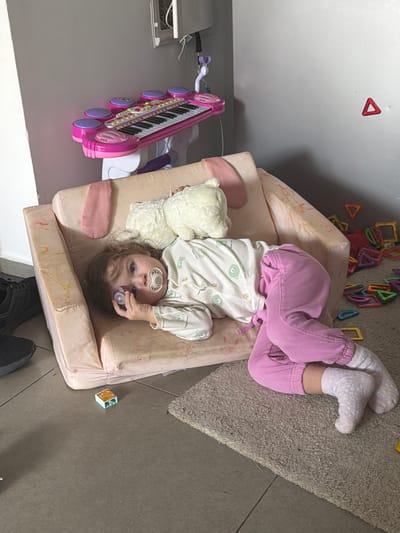 A child lies on a soft, peach-colored chair with a stuffed teddy bear. The child wears pink pants and a patterned pajama top. A pacifier is in their mouth. The child is surrounded by colorful toys including magnetic tiles and a pink toy piano. The scene takes place indoors. The location is likely Harish, Israel.