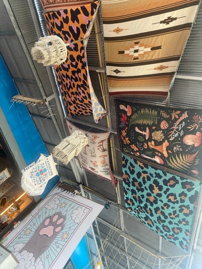 The image, taken from a low angle, shows the ceiling of a building, possibly a market or fair, in Baan Nai Suan, Thailand. The ceiling is made of corrugated metal. A series of tapestries or blankets are suspended horizontally across the ceiling, creating a decorative effect. The tapestries feature various patterns: a striped design in earthy tones, a dark tapestry with botanical illustrations including mushrooms, a teal tapestry with a leopard print, and an orange tapestry with a similar leopard print. Underneath the tapestries hang decorative items, including macrame-style light fixtures and banners displaying the text "#XXCULTURE THE WORLD AHEAD" along with illustrations. In the lower right corner, a large poster depicting a cartoon cat paw surrounded by clouds and stars is visible. The overall scene suggests a bohemian or artistic aesthetic.