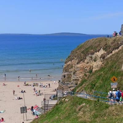 The image captures a coastal scene at Ballybunnion, Ireland, on a clear, sunny day. In the foreground, a steep, grassy cliff face dominates the right side of the frame, with a paved pathway and a blue railing following its contour, indicating a scenic walking route. A "CAUTION" sign with a falling rocks symbol and a "no swimming" symbol is visible on the cliff face.
Below the cliff, a sandy beach stretches out, populated by numerous beachgoers. People are seen sunbathing on towels, building sandcastles, wading in the shallow waters of the ocean, and playing near the shoreline. The ocean itself is a vibrant blue, with gentle waves lapping at the shore. In the distance, a long, low-lying island or peninsula is visible on the horizon under a clear blue sky.
Several people are walking along the path on the cliff, descending towards the beach. One person is seen kayaking further out in the sea. The scene overall conveys a typical summer day at a popular seaside location.
