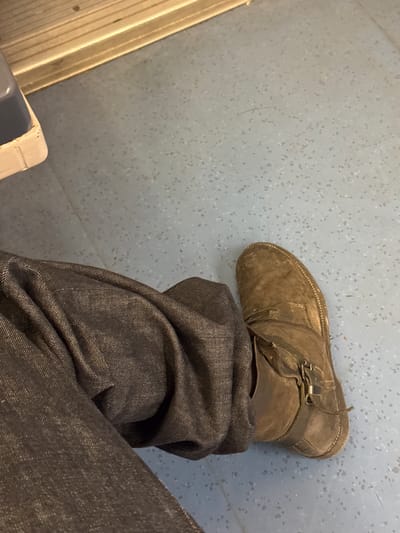The image captures a close-up, top-down view of a person's lower leg and foot within an indoor setting, likely public transport. The primary subjects are a dark, possibly olive-green or brown, worn boot with a strap and buckle detail around the ankle, and dark-colored, likely denim, pants that are creased and bunched up over the boot. The boot's material appears to be suede or a similar textured fabric.

The scene is set on a light blue-grey floor with a distinct speckled or flecked pattern, commonly found in buses, trains, or metro cars. Along the far left edge of the frame, a metallic or light-colored rail or track is visible, further suggesting a public vehicle interior. In the bottom-left corner, a small fragment of what appears to be a seat's base or armrest, featuring a light and dark blue component, can be discerned.

The person is stationary, with their foot resting on the floor. No specific activity or event beyond being present in this space is depicted. The image does not provide cues for time of day or weather. The setting aligns with public transportation in an area like Palaiseau, France. There is no visible text within the frame.