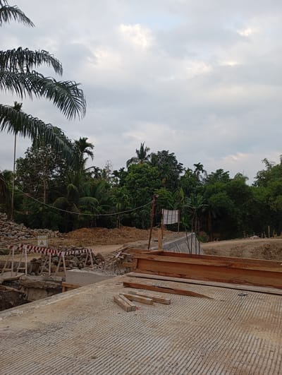 In Blangraleu, Indonesia, a construction site features a partly finished concrete structure in the foreground, marked with parallel grooves from its creation. Wooden planks lie scattered on the concrete surface, and a large wooden formwork is positioned to the right. Beyond this, a damaged bridge or culvert is visible, with rocks and debris piled up. A striped barrier with wooden supports blocks the immediate area. A yellow sign with a black triangle warning symbol is placed near the rocks. In the background, a dense array of tropical trees and foliage, including palm trees and bamboo, forms a lush green canopy against a cloudy sky.