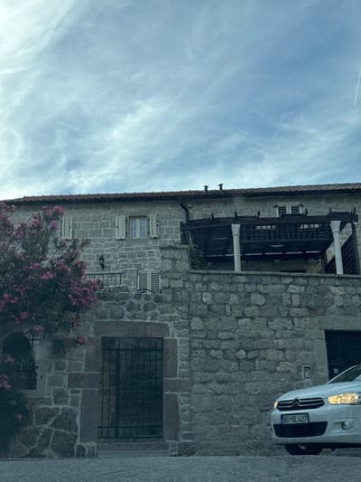 In the city of Rijeka Reževići, Montenegro, a white car with license plate BO BL 427 is parked in front of a rustic stone building. The car is facing towards the right of the frame, with its headlights on. The building features stone walls, wooden shutters on the windows, and a tiled roof. A flowering bush with pink blossoms is visible to the left of the building. The sky above is overcast with wispy clouds.