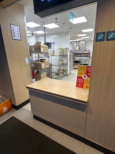 This image depicts the interior of a commercial food service establishment, specifically a delivery or takeaway pickup point and its adjoining kitchen area, located in Chatham, United Kingdom.
In the foreground, a light wood-effect counter, approximately waist-height, extends horizontally across the frame. Its top edge features a thin dark strip. On the right side of this counter, a red and yellow "McDelivery" freestanding sign is angled towards the viewer, displaying text and icons related to delivery services. The floor in front of the counter is tiled in a light, neutral color, partially covered by a dark rectangular floor mat on the left. An orange "Just Eat" thermal delivery bag rests on the floor to the far left, leaning against a light wood-paneled wall section. This wall section also features a framed sign depicting packaging rules (checked and crossed items).
Through an open archway above and behind the counter, a brightly lit commercial kitchen environment is visible. The ceiling is white with multiple square recessed lighting panels and some hanging fixtures. Green emergency exit signs and small black security cameras are mounted on the ceiling. A large black digital display screen hangs directly above the archway, showing partial text like "0 C" and "Order."
Within the kitchen, to the left, a metal shelving unit is stocked with numerous brown cardboard boxes, some open, suggesting inventory or prepared items. A partially obscured individual, appearing to be a staff member, is visible behind this shelving unit. To the right of the shelving unit, another multi-tiered metal cart or shelving unit holds stacks of paper or plastic containers. Further into the background, the kitchen features stainless steel commercial equipment, including hot holding units or fryers, and a visible digital order screen above a counter. The kitchen floor is tiled in a light color, distinct from the darker floor mat in the foreground. Wall surfaces in the kitchen are predominantly white or light-colored. Two square "Deliveroo" logos, featuring a wireless signal icon, are affixed to the wooden wall panel on the far right, near the archway opening.