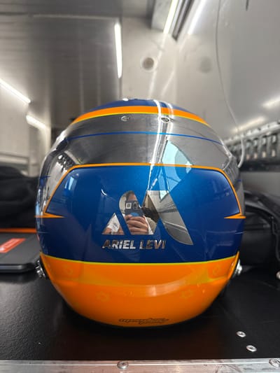 The image shows a racing helmet lying on a metal surface. The helmet is blue and orange, with a silver top. The helmet has the words "Ariel Levi" written on the side in silver letters. There is a reflection of a person in the helmet's visor, holding a phone. The helmet is likely located in a garage or workshop, as evidenced by the lighting and surrounding objects. The coordinates provided (latitude: 41.57, longitude: 2.259) correspond to a location near the Circuit de Barcelona-Catalunya in Montmeló, Spain. This suggests that the helmet belongs to a racer or member of a racing team.
