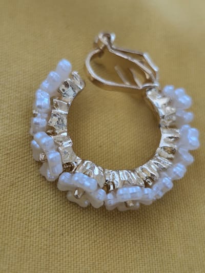 The image shows a close-up, eye-level, outdoor shot of a single, gold-colored hoop earring with white beads, resting on a textured yellow surface. The earring is partially obscured by the texture of the surface, which appears to be fabric. The earring is made of a circular gold metal base, with a textured, slightly irregular surface. Small, white, seed-like beads are attached to the outer edge of the earring, forming a decorative border. The earring has a hinged clasp mechanism on the top portion of the hoop. The background is a consistent yellow-brown color with visible texture, suggesting a tablecloth or similar fabric. The lighting appears to be natural, casting subtle shadows on the earring. No people, animals, or identifiable landmarks of Fresnes, France are present in the image.