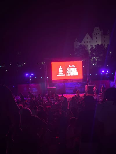 The image shows a crowd of people at an outdoor event at night in Ingolstadt, Germany. A large, bright red screen displays a logo and text, likely for a sponsor or event branding. Behind the screen, a pale-colored building, possibly a castle or historic structure, is visible against the dark sky, illuminated by stage lights. The crowd is facing the screen, suggesting a performance or presentation is taking place. The lighting is predominantly purple and red, characteristic of nighttime events. The atmosphere appears energetic, with many people gathered. Some blue spotlights are also visible in the background. The time of day is clearly night. The text on the red screen is partially visible and includes the numbers "2020" and "2027", along with what appears to be a stylized logo and the word "Sparkasse", a German financial institution.
