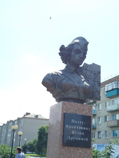 The image shows a bust of a woman wearing a military-style cap and holding a book or scroll. The bust is made of dark metal and is mounted on a granite pedestal. A plaque on the pedestal reads "Поэту-фронтовику Юлии Друниной" (To the front-line poet Yulia Drunina). The woman's expression is serious. In the background, there are apartment buildings, trees, and streetlights. A bird is visible in the sky. The overall scene appears to be an outdoor public space in the city of Yalutorovsk, Russia, likely a park or square.