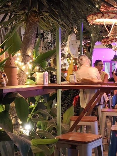 The image shows an outdoor bar or restaurant scene in Rotes, Spain. In the foreground, there is a wooden shelf or table decorated with a skull, a small tower-shaped ceramic item, a candle in a glass holder, and other small objects. Behind the table, lush tropical foliage, including large leaves, surrounds a tree trunk adorned with fairy lights. In the background, two women are seated at a higher table. The woman on the left has gray hair pulled back in a bun, and the woman on the right wears glasses and a yellow top. They appear to be engaged in conversation. There is a fan on a pole, and colorful lanterns hang overhead. The lighting is soft, with a mix of artificial light from the lanterns and the fairy lights. The overall setting is relaxed and tropical.